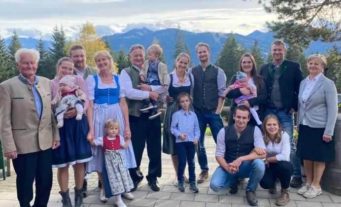 Drei Generationen einer Familie in Trachtenkleidung vor einer Bergkulisse, freundlich lächelnd.